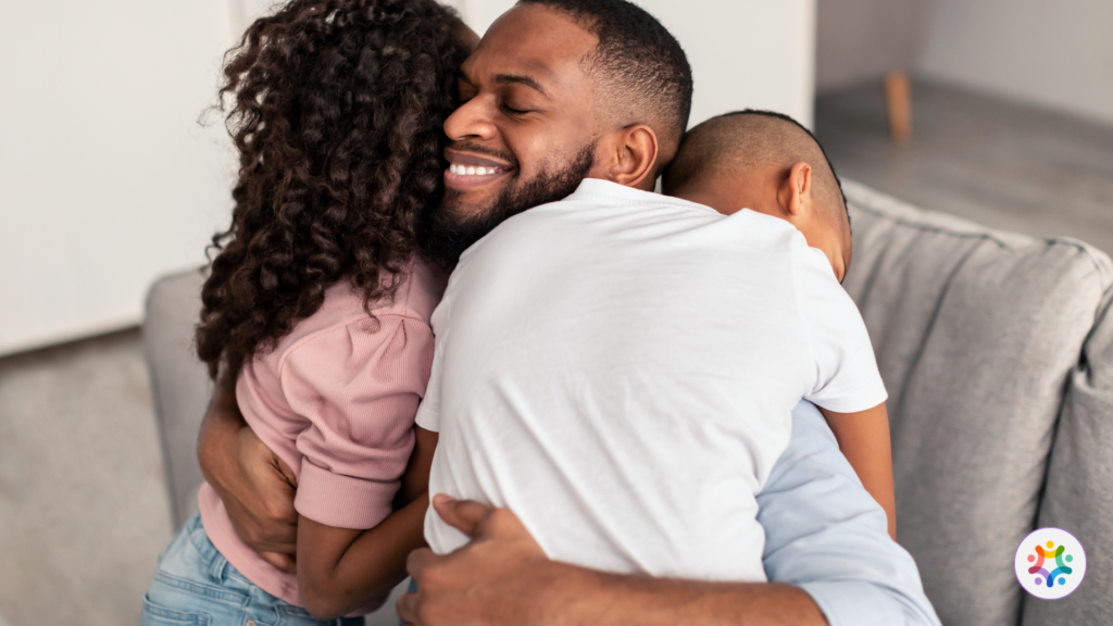 Foto de um pai sentado no sofá, corrindo, abraçando sua filha e seu filho.